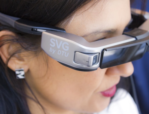 A person is wearing a pair of advanced augmented reality glasses with the label 'SVG by DTU' on the side, indicating a technological product. The glasses have a sleek, modern design and cover the eyes of the individual. The person has dark hair and is seen in a close-up view, focusing primarily on the device.